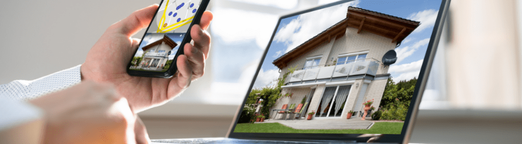A hand holding a phone with an image of a real estate listing in front of a computer screen with a house on it